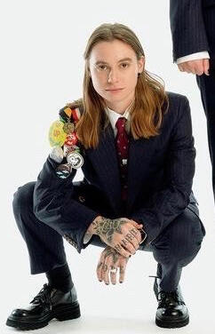 picture of julien baker, a twentysomething year old white woman with light brown hair, dark eyes, and tattoos. she is dressed in a black suit with colorful pins and is squatting.