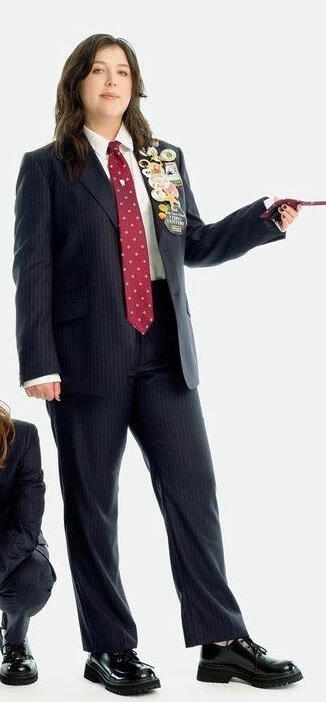 picture of lucy dacus, a twentysomething year old white woman with dark brown hair and dark eyes. she is dressed in a black suit with colorful pins and is standing holding a tie offscreen.
