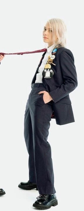 picture of phoebe bridgers, a twentysomething year old white woman with bleached silver hair and dark eyes. she is dressed in a black suit with colorful pins and is standing with her tie held offscreen.